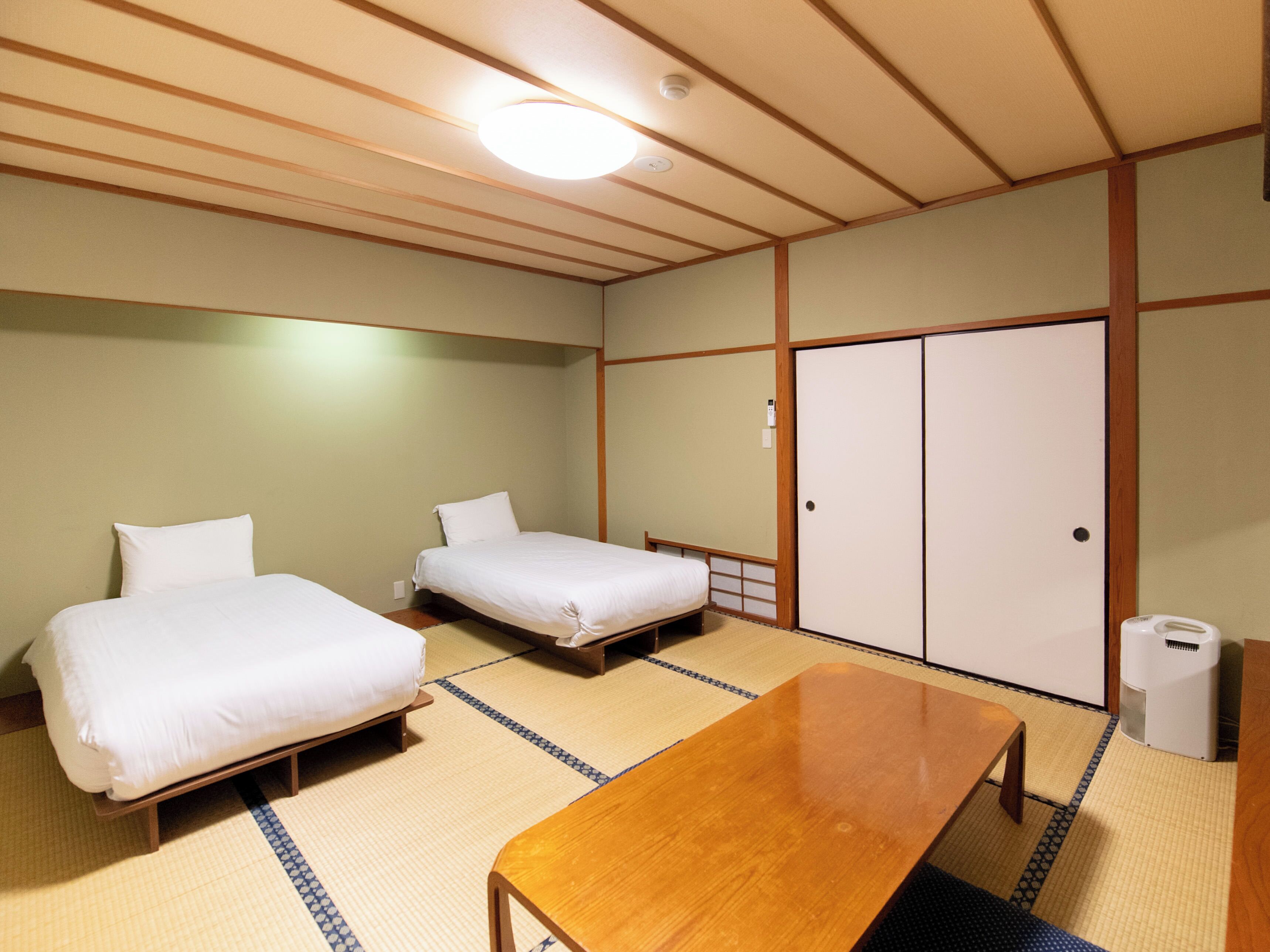 A room with two beds, a wooden table, tatami flooring, and a wardrobe.