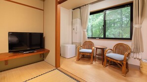 Una habitación con televisión, dos sillas de mimbre, una mesita, refrigerador y ventana con vista a árboles.