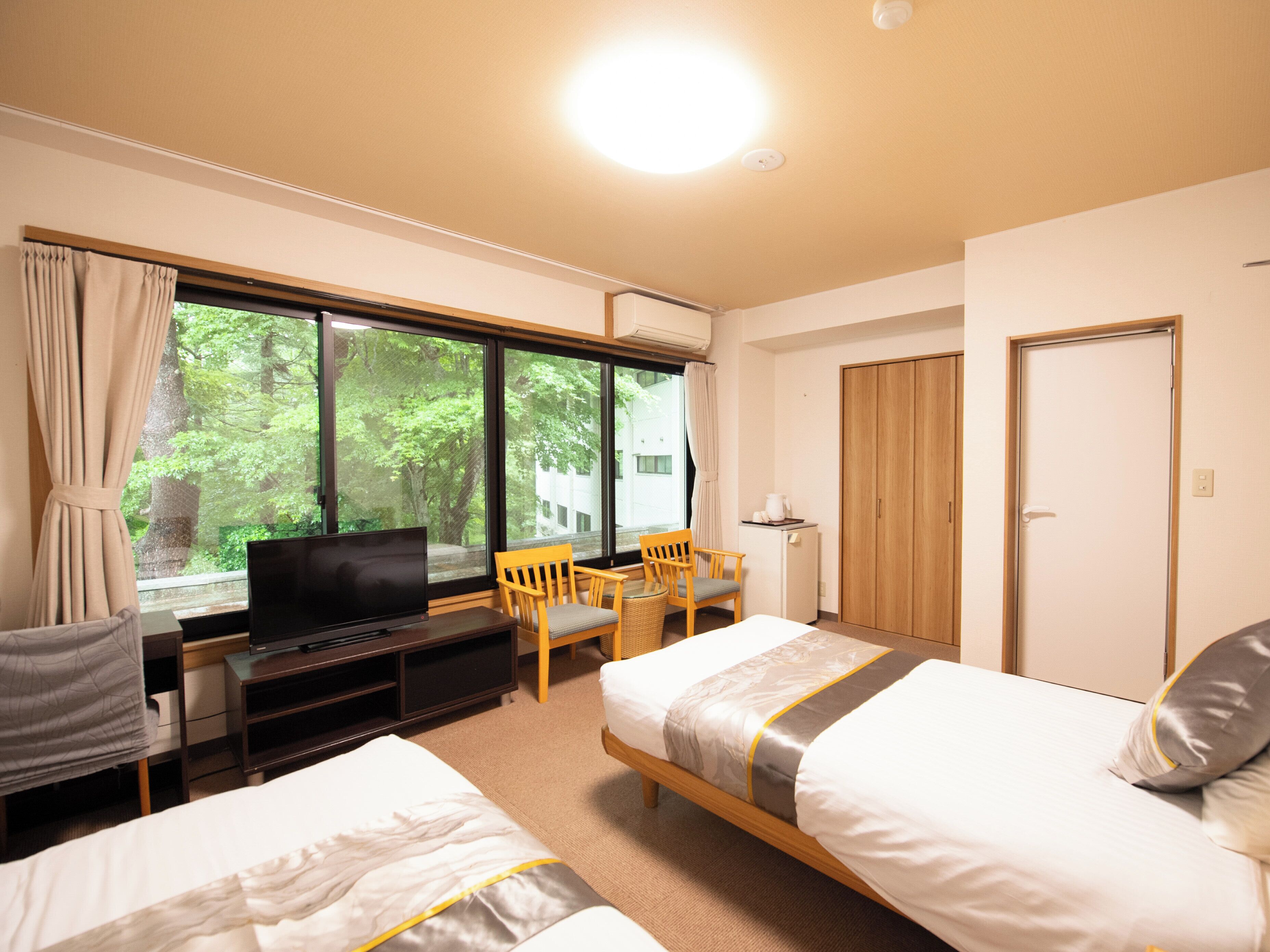Ein Hotelzimmer mit zwei Betten, einem Schreibtisch mit Fernseher, einem Sessel, einem Kleiderschrank und einem Fenster mit Blick auf Bäume.