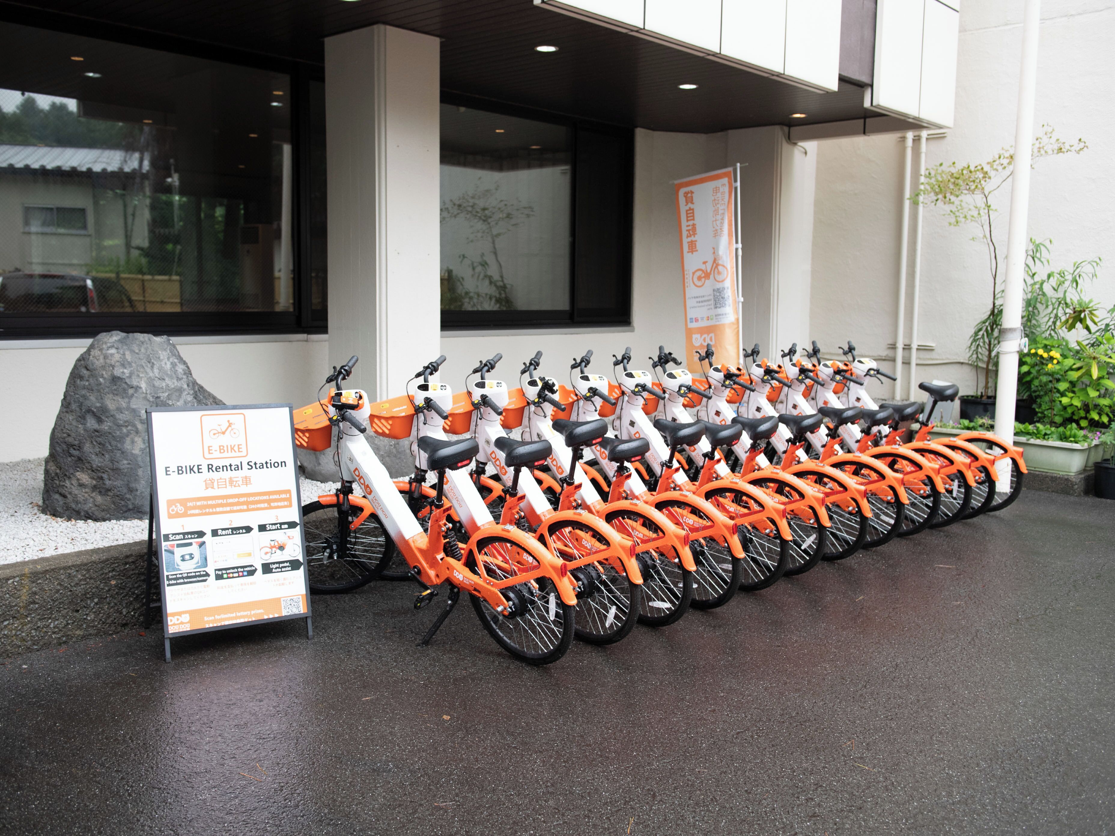 Une rangée de vélos électriques orange est stationnée devant un bâtiment sur lequel est apposée une enseigne indiquant qu’il s’agit d’un point de location.