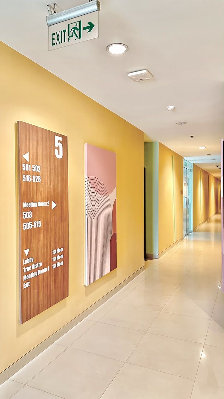 A hotel corridor with directional signs, a wooden panel with room numbers, and an exit sign.
