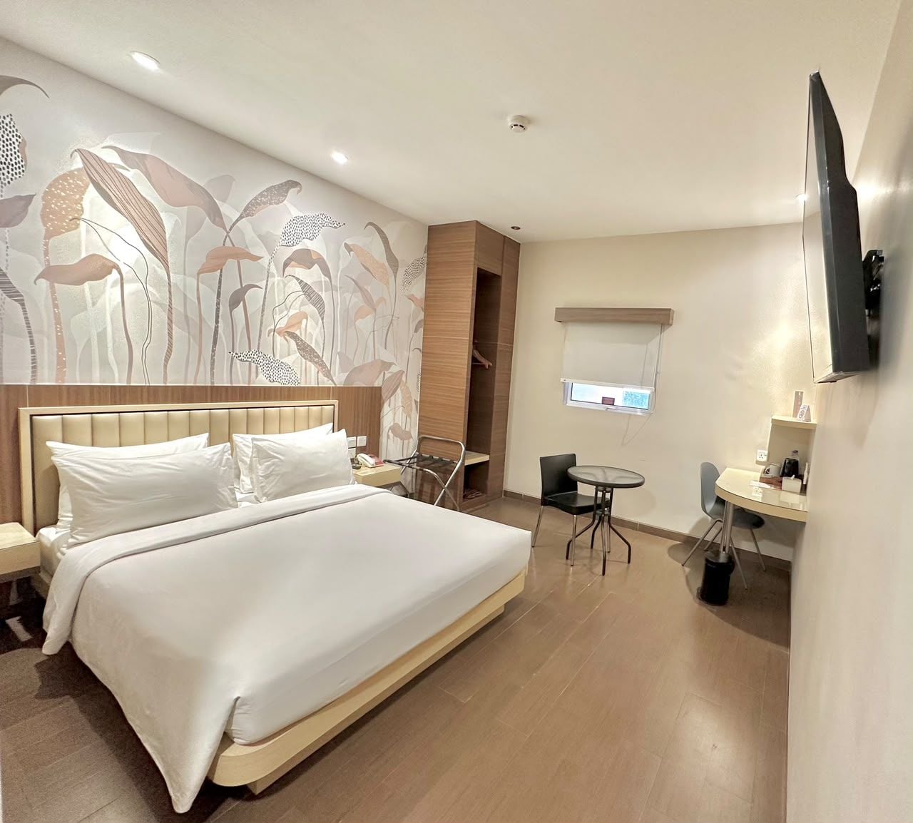 Quarto de hotel com uma cama grande, escrivaninha, cadeira, televisão e parede com padrão floral.