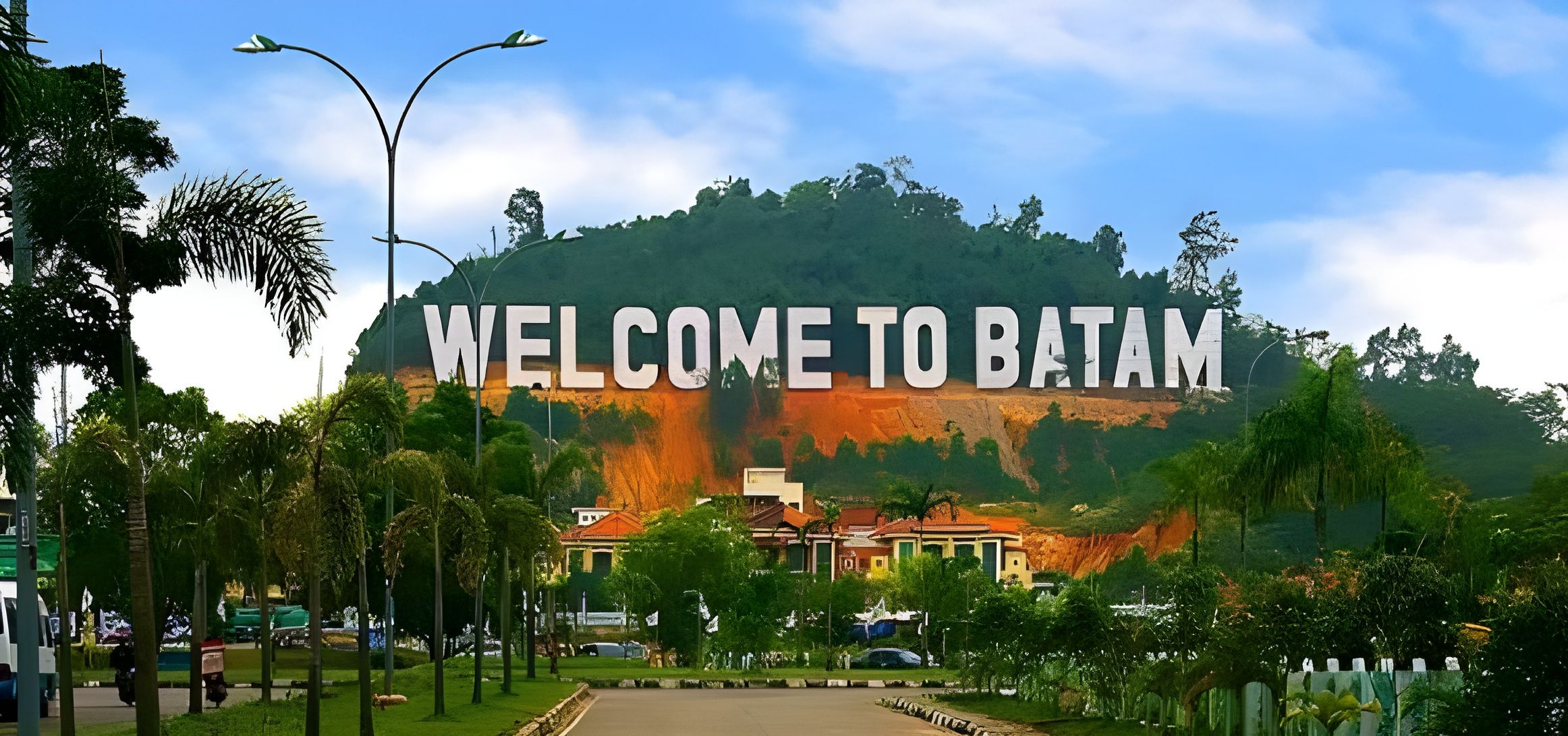 En velkomstskilt med 'WELCOME TO BATAM' foran en frodig grøn bakke og et boligområde med huse og træer.