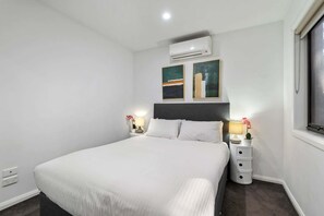 A bedroom with a bed, two bedside tables with lamps, an air conditioning unit, and two abstract paintings on the wall. - Comfortable 2BR Retreat with Balcony & Parking (St Kilda)
