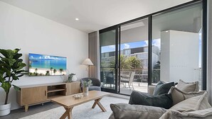 A modern living room with a flat-screen TV, a wooden coffee table, and a sofa with cushions. There is a large window offering a view of a balcony with outdoor furniture.