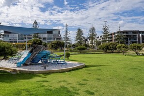 A playground with slides and seating, surrounded by green grass and trees, with modern buildings in the background. - Trendy 1-Bed Retreat in Fremantle with Rooftop (North Fremantle)