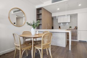 A modern kitchen with white cabinets, a wooden dining table with wicker chairs, and a round mirror on the wall. - Trendy 1-Bed Retreat in Fremantle with Rooftop (North Fremantle)