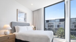 Een moderne hotelkamer met een groot bed, twee nachtkastjes met lampen, een kunstwerk in een lijst boven het bed, en een groot raam met uitzicht op de stad.