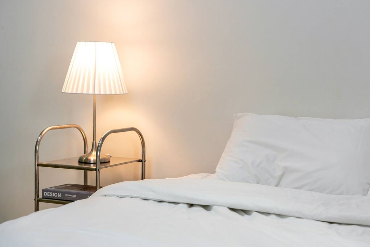 Ein ordentlich gemachtes Bett mit weißer Bettwäsche, eine Nachttischlampe und ein Metall-Nachttisch mit einem Buch mit dem Titel „DESIGN“.