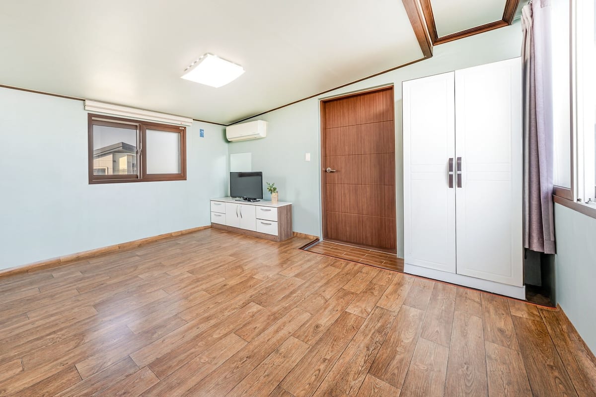 Ein geräumiges Zimmer mit Holzboden, einem weißen Kleiderschrank, einer kleinen Anrichte mit Fernseher und einem Fenster mit Blick auf ein Gebäude.