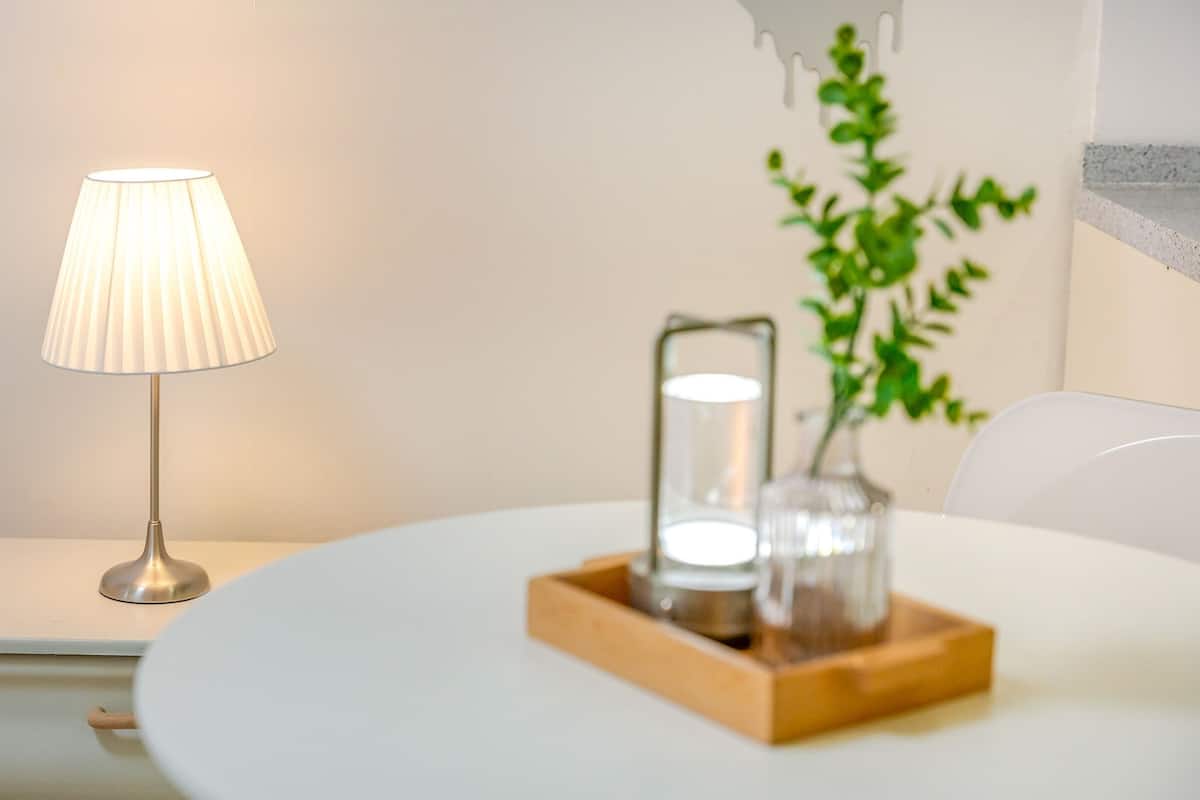 Ein ordentlich eingerichteter Raum mit Nachttisch, Lampe und einem kleinen Holftischchen, auf dem ein Einmachglas und eine Pflanze stehen.