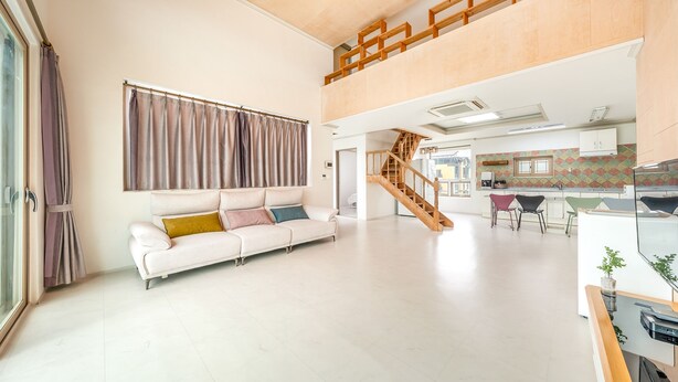 Un amplio salón con un sofá blanco, una escalera que lleva a un nivel superior, y una zona de cocina con azulejos de colores.