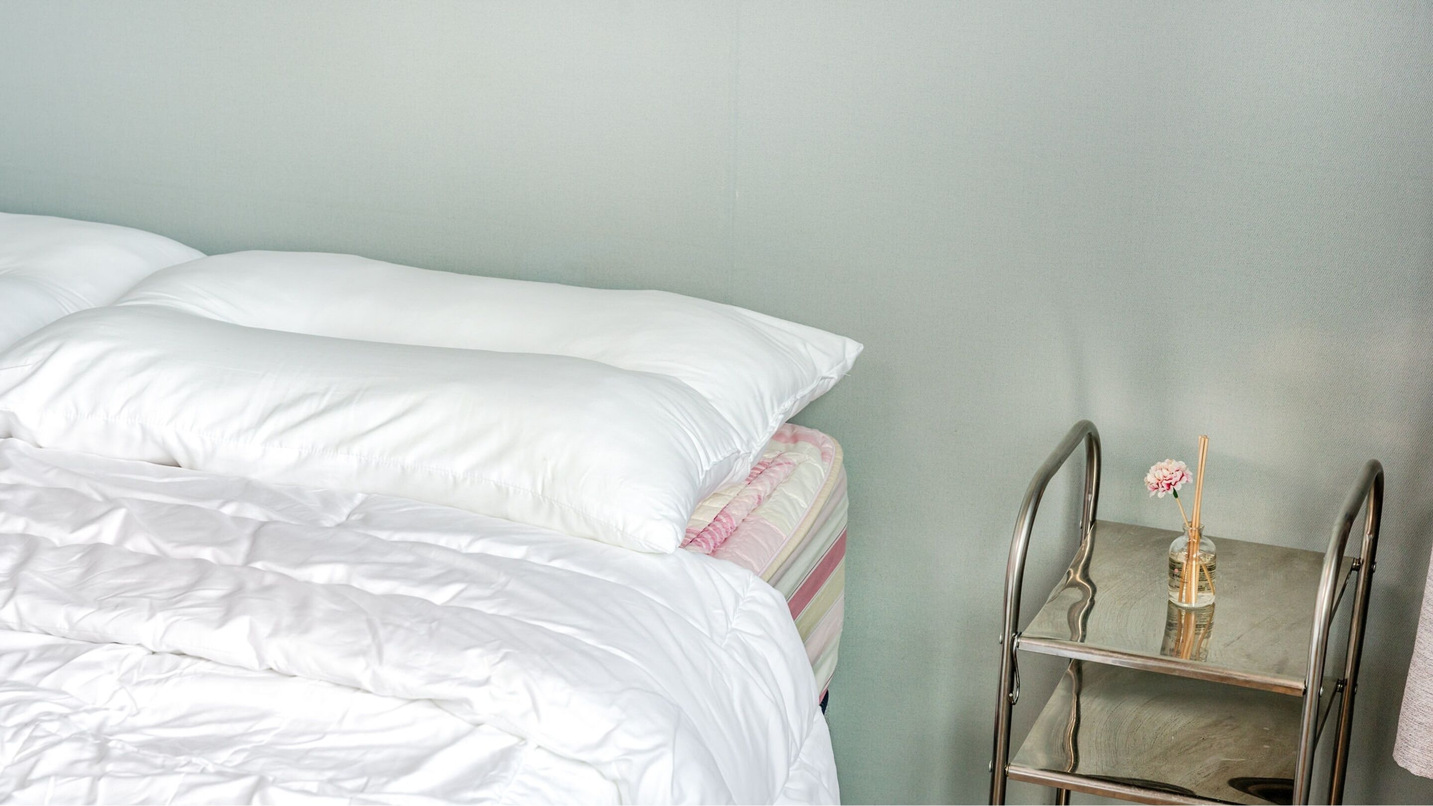 A neatly made bed with white bedding, a metal bedside table with a glass container and a pink flower, and a light green wall.
