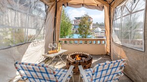 Una carpa con un brasero y dos sillas, una mesa con una botella y vasos, y vistas a edificios y árboles.
