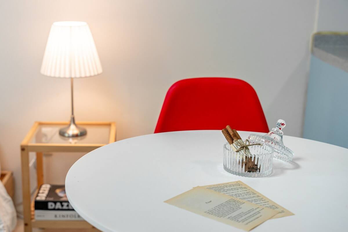 Ein ordentlich eingerichtetes Hotelzimmer mit einem weißen runden Tisch, einem roten Sessel, einer Nachttischlampe und einem kleinen Nachttisch mit Büchern.