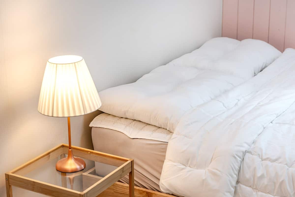 Ein ordentlich bezogenes Bett mit weißer Daunendecke und Kissen, ein Nachttisch mit Lampe und ein Holzbeistelltisch.