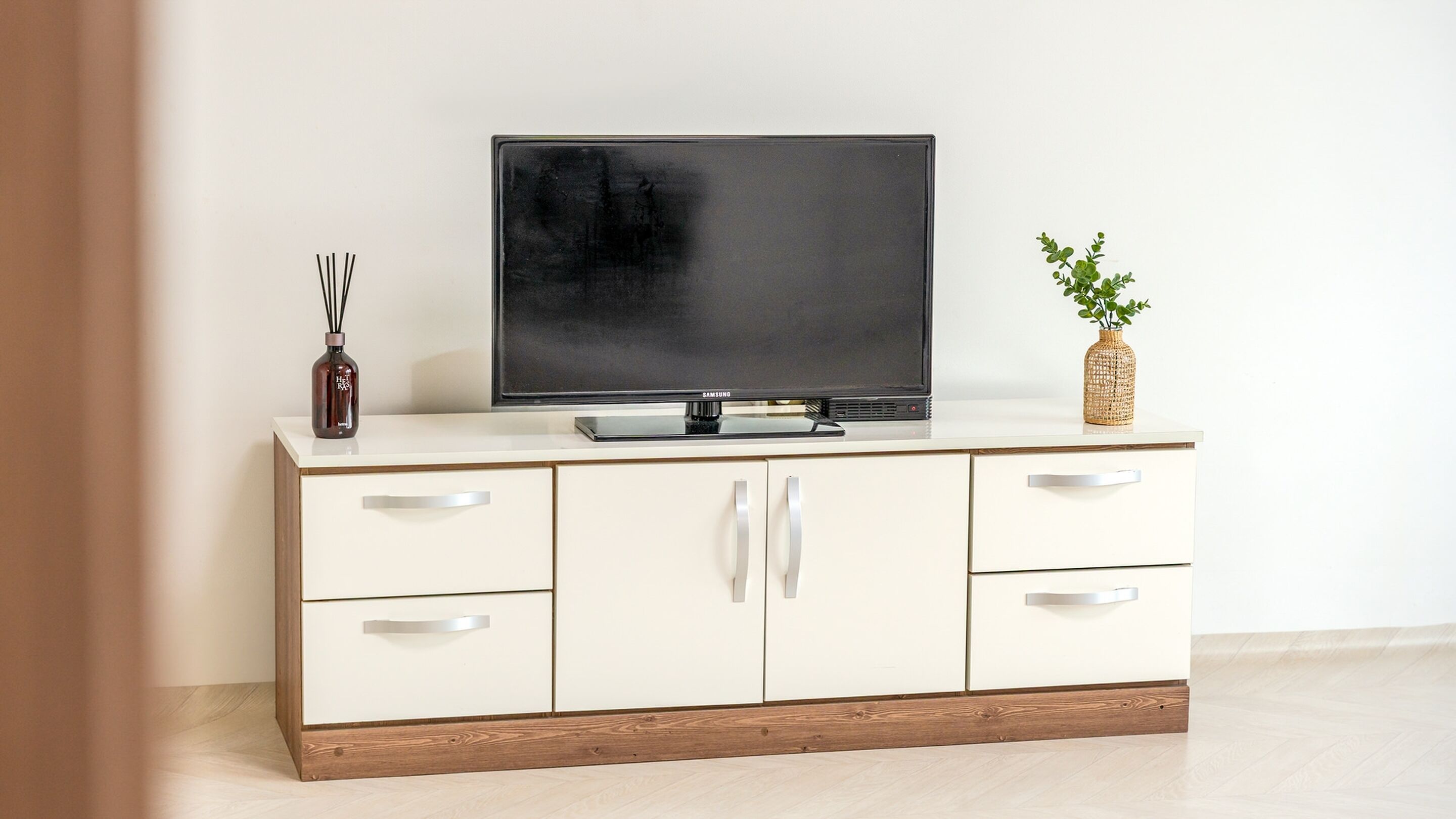 A white cabinet with a flat-screen TV on top, a small vase with green plants, and a bottle of reed diffuser.