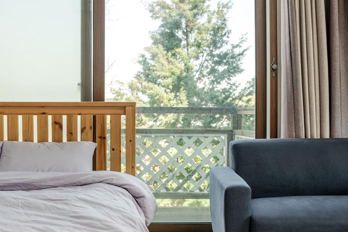 Ein Schlafzimmer mit einem hölzernen Kopfteil, einem Bett mit weißer Bettwäsche, einem grauen Sessel und Blick auf Bäume durch ein Fenster mit Vorhängen.