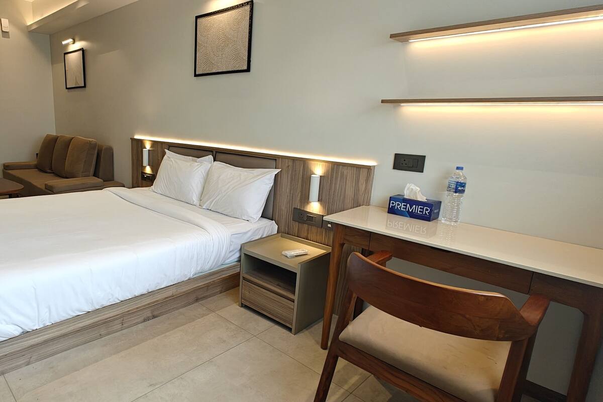 A hotel room with a bed, a desk, a chair, a nightstand, a sofa, and a kitchenette.