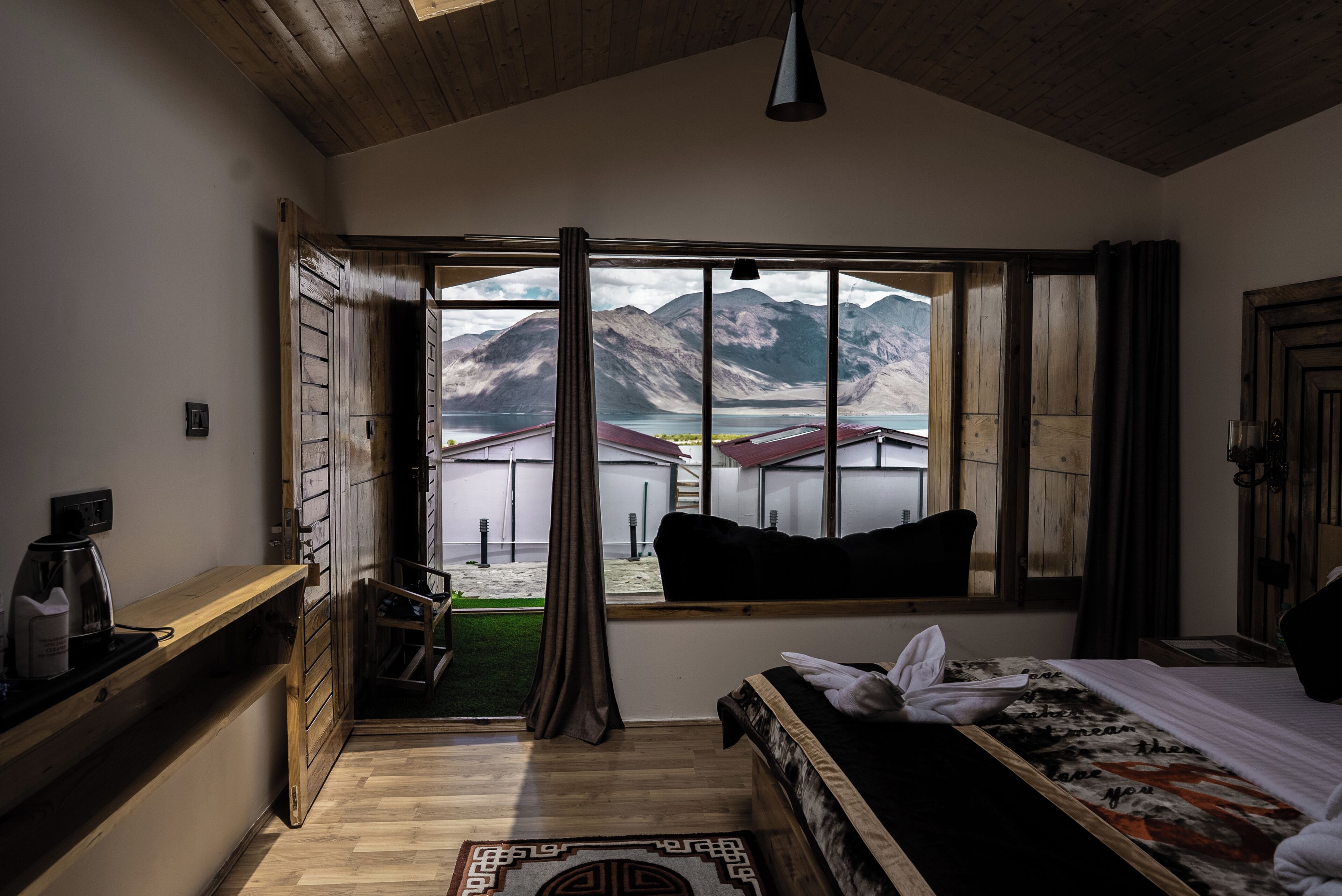 A room with a large window, wooden ceiling, a bed, a bench, and a small table with a kettle.