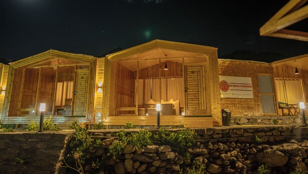 Een houten chalet-achtig gebouw 's nachts, met een verlichte ingang, een bord en een stenen muur op de voorgrond.
