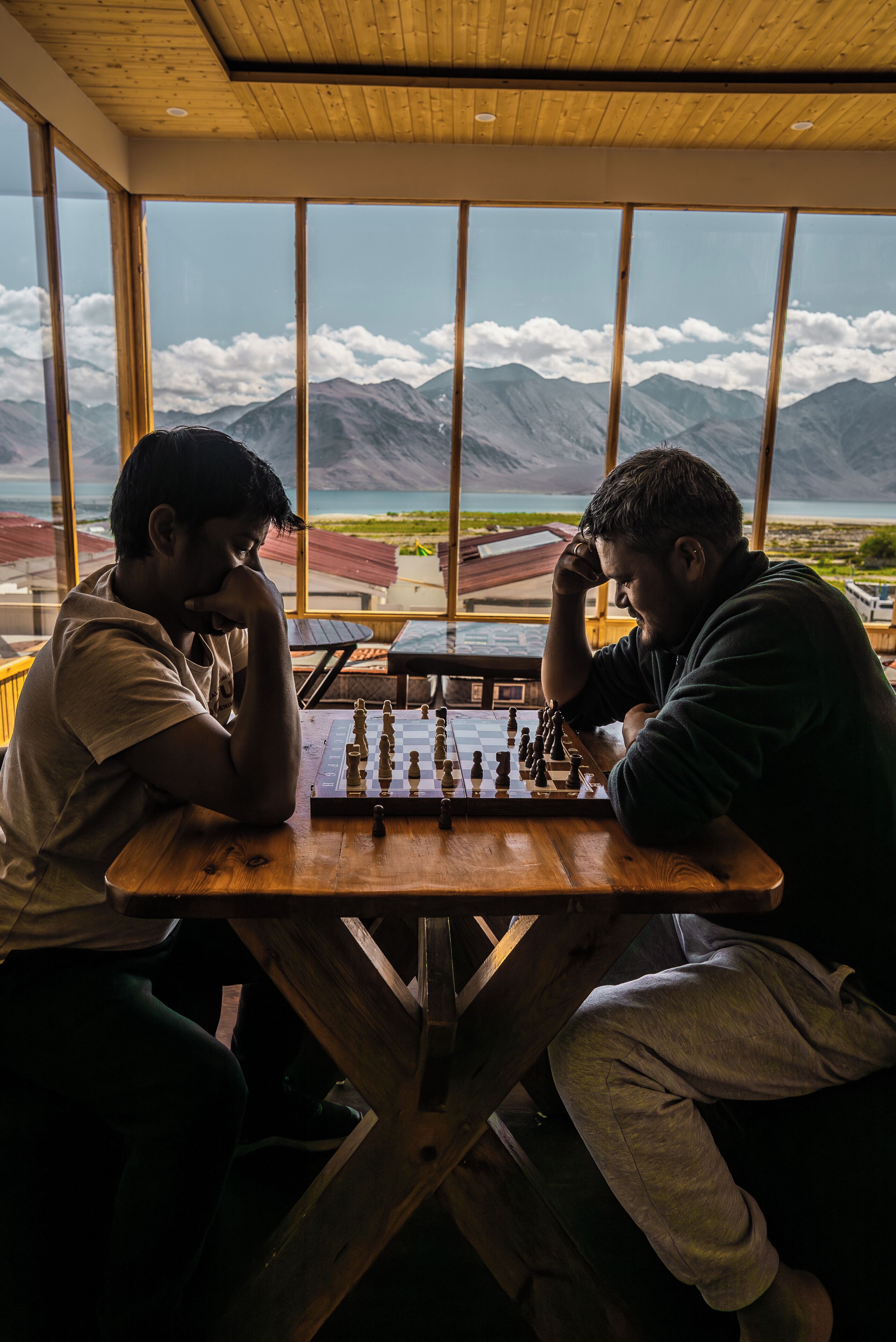 Zwei Personen spielen Schach an einem Holztisch, mit Blick auf eine Berglandschaft durch große Fenster.