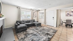 Una sala de estar con un sofá seccional, una chimenea, una zona de comedor con mesa y sillas, y un ventanal con cortinas.