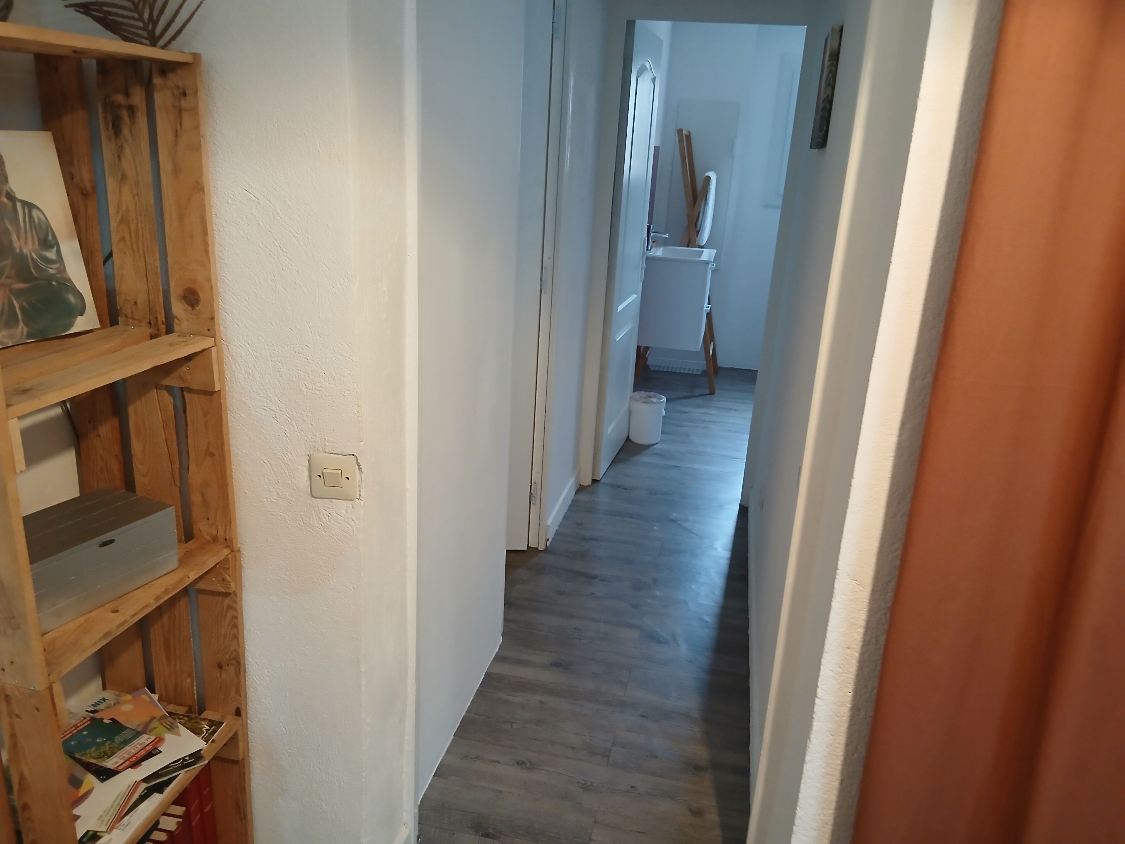 A hallway with a wooden shelf unit on the left, a white wall, and a wooden floor.