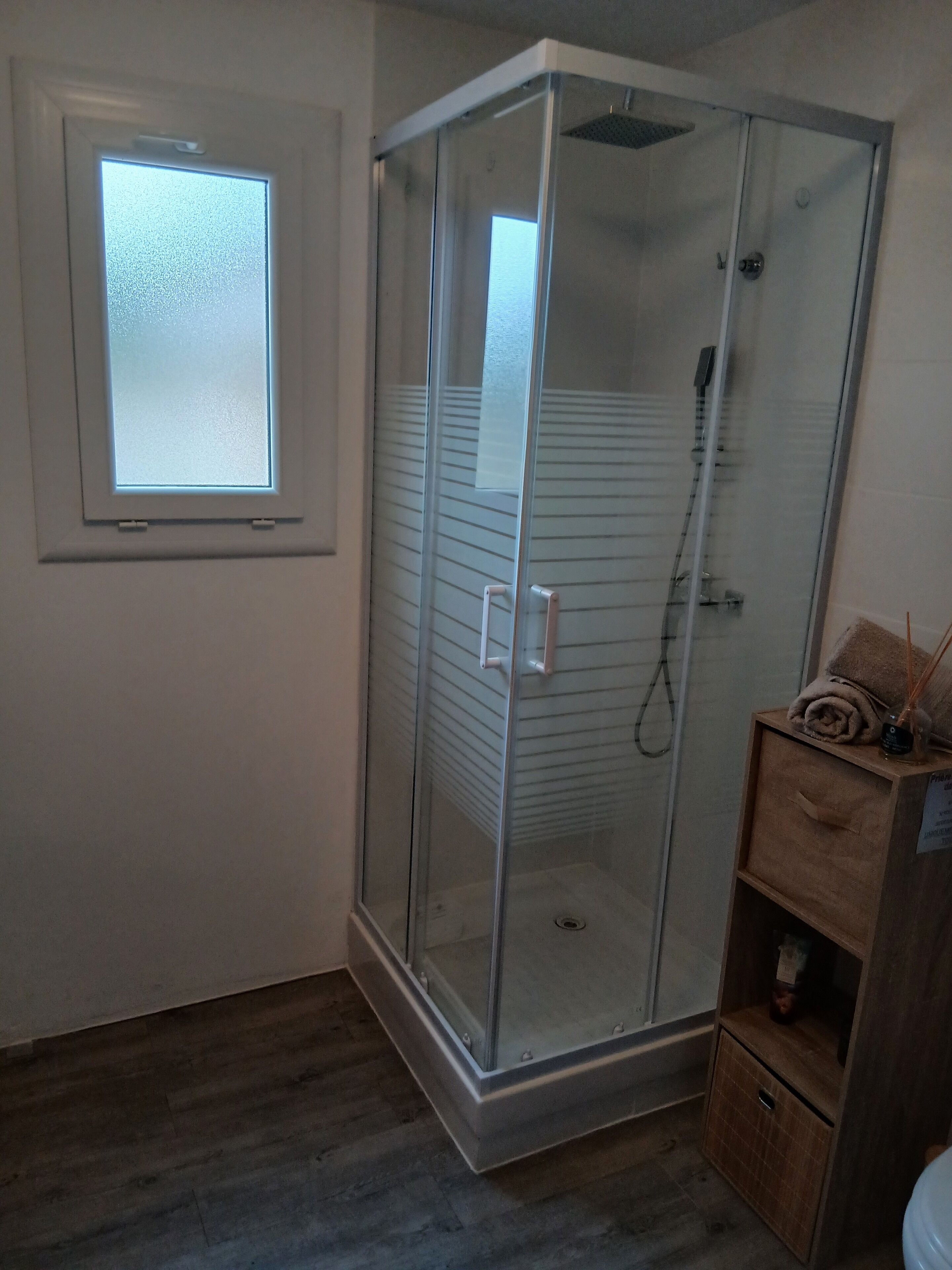 A glass-enclosed shower with a handheld showerhead, a small wooden shelf with a towel and a bottle, and a window with frosted glass.