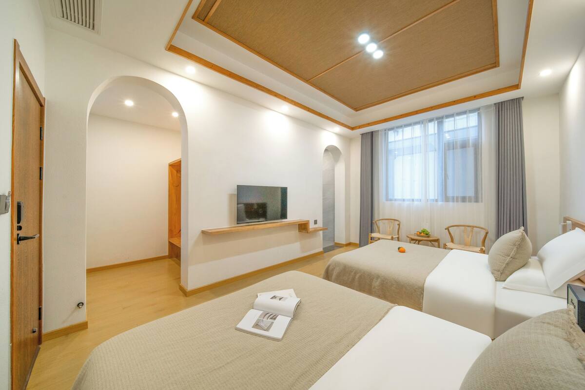 A hotel room with two beds, a TV, a small table with a fruit, and a window with curtains.