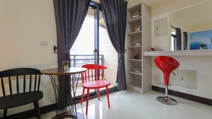 A room with a dining table, two chairs, a red chair, a red stool, a shelf with decorative items, and a window with curtains. - XIA XIA JIAO HOMESTAY (Magong)