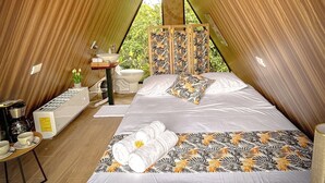 A cozy, triangular-roofed bedroom with a large bed, a bedside table with a kettle, and a view of greenery outside. - Tilawa La Fortuna Glamping (La Fortuna)