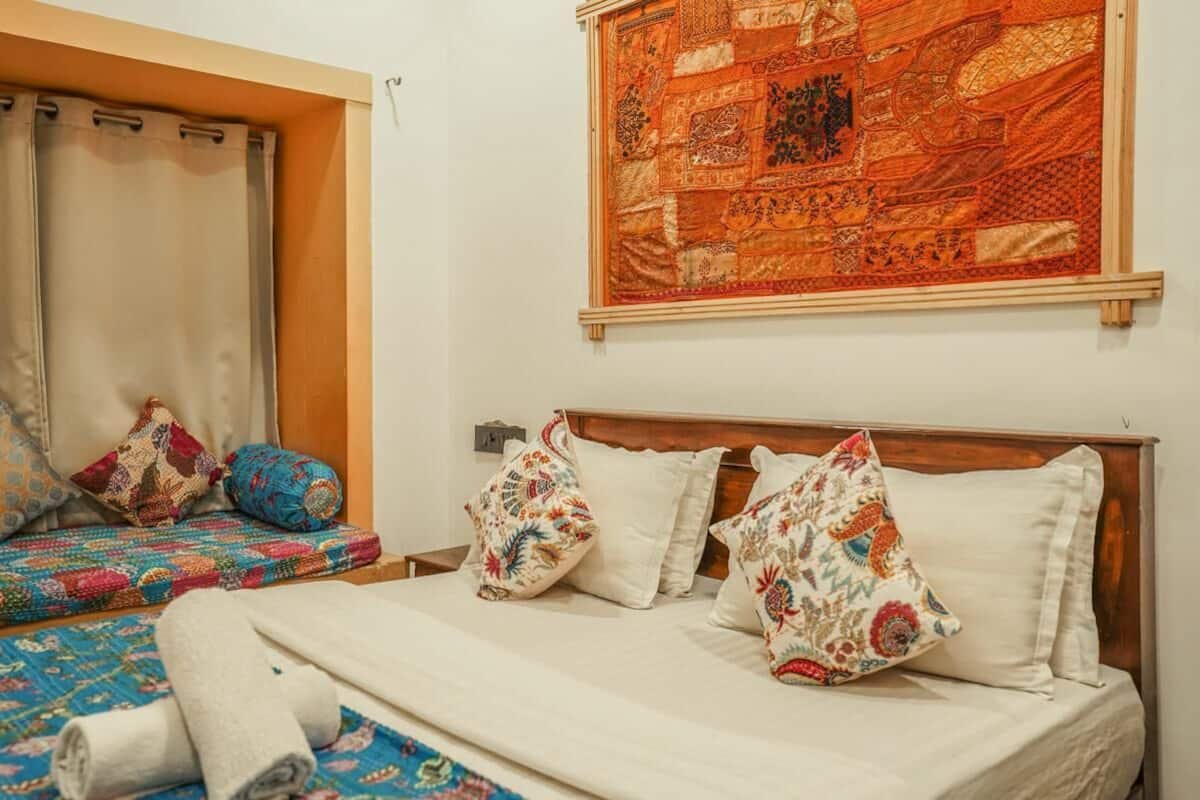 A bedroom with a bed, pillows, a quilt, a window with curtains, and a tapestry on the wall.
