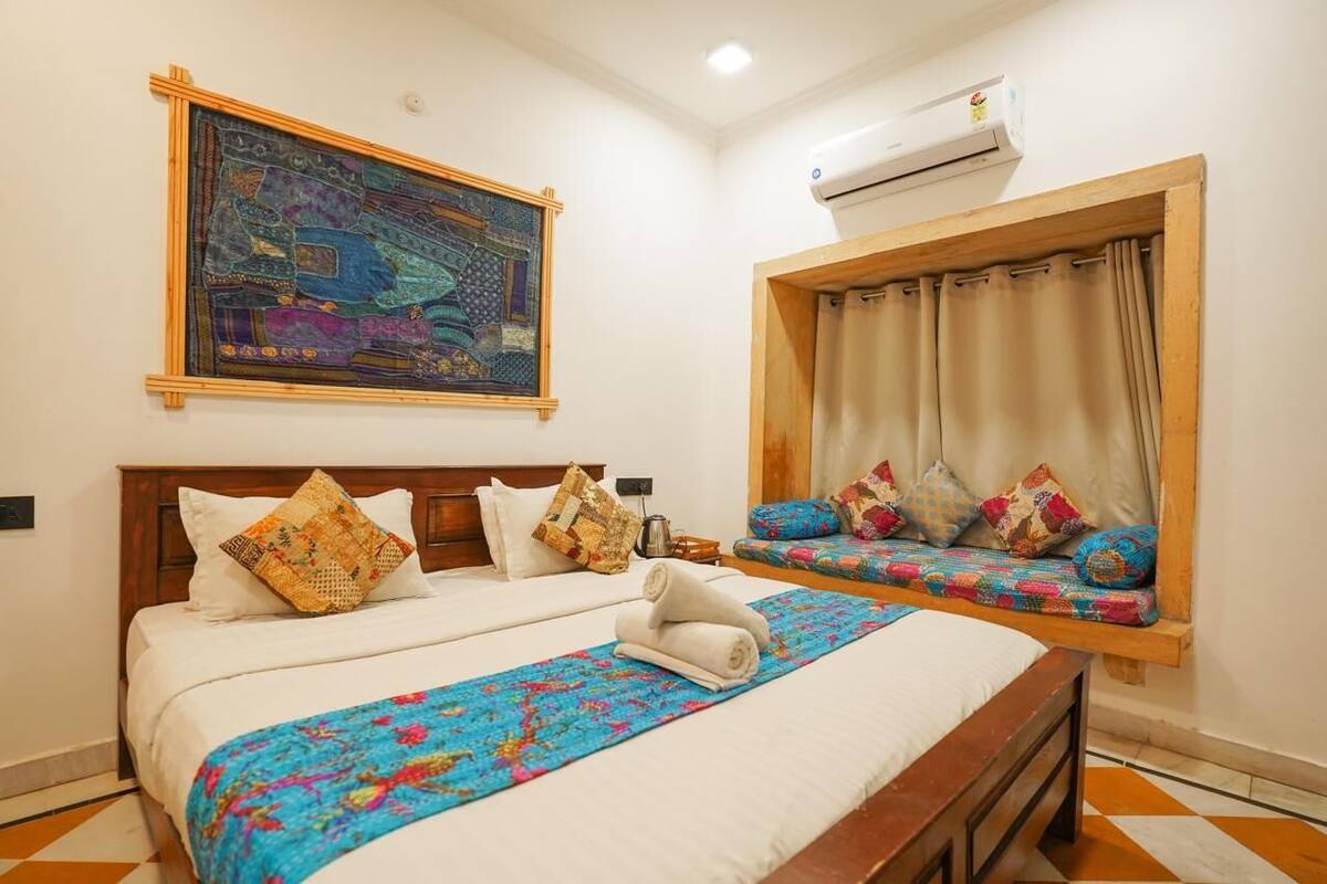 A hotel room with a large bed, a smaller sofa bed, a colorful artwork on the wall, and an air conditioning unit.