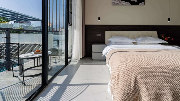 Une chambre moderne avec un grand lit, une table de chevet et une porte vitrée donnant sur un balcon équipé d’une chaise et d’une table.