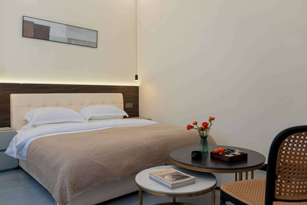 A neatly made bed with a headboard, a bedside table with a book, a small round table with a vase of flowers, and a wicker chair.