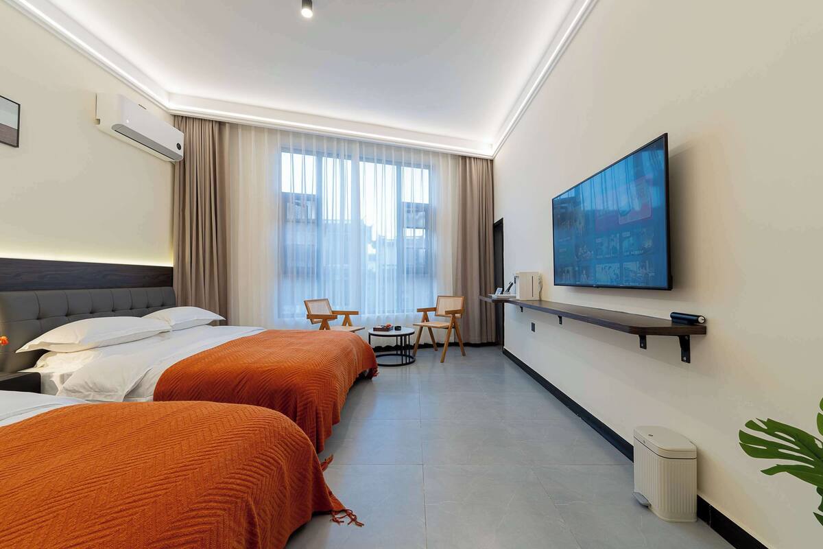 A hotel room with two beds, a TV, a small table, chairs, and a window with curtains.