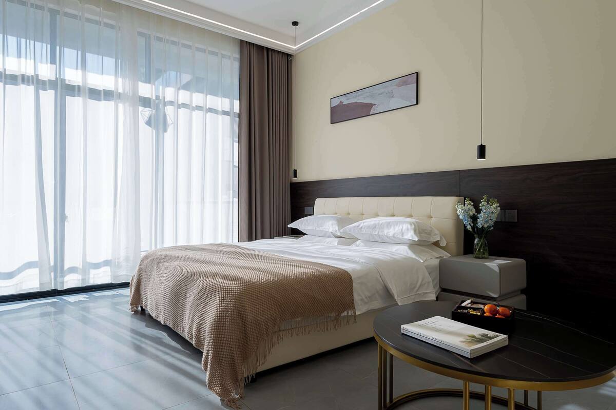 A modern hotel room with a large bed, a bedside table with a book and fruit, a dark headboard, and a large window with sheer curtains.
