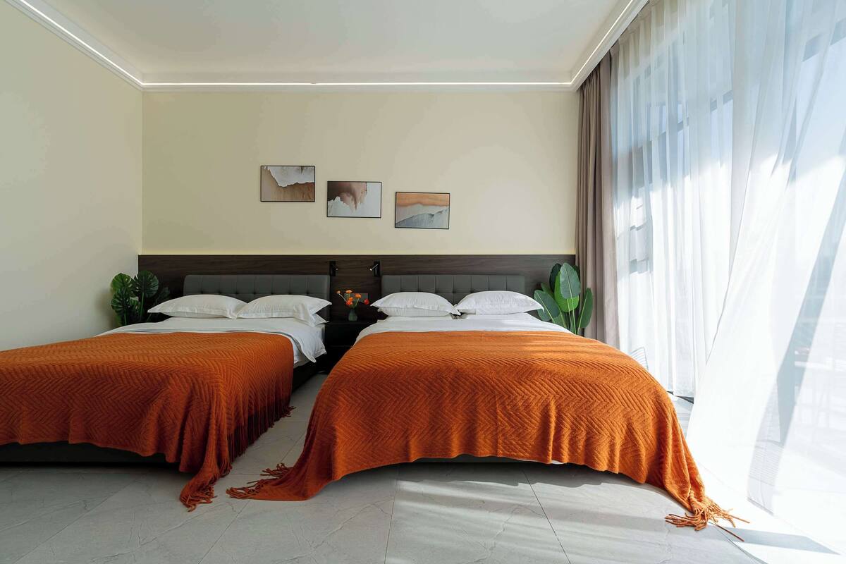 A hotel room with two beds, a large window with sheer curtains, and three wall-mounted paintings.