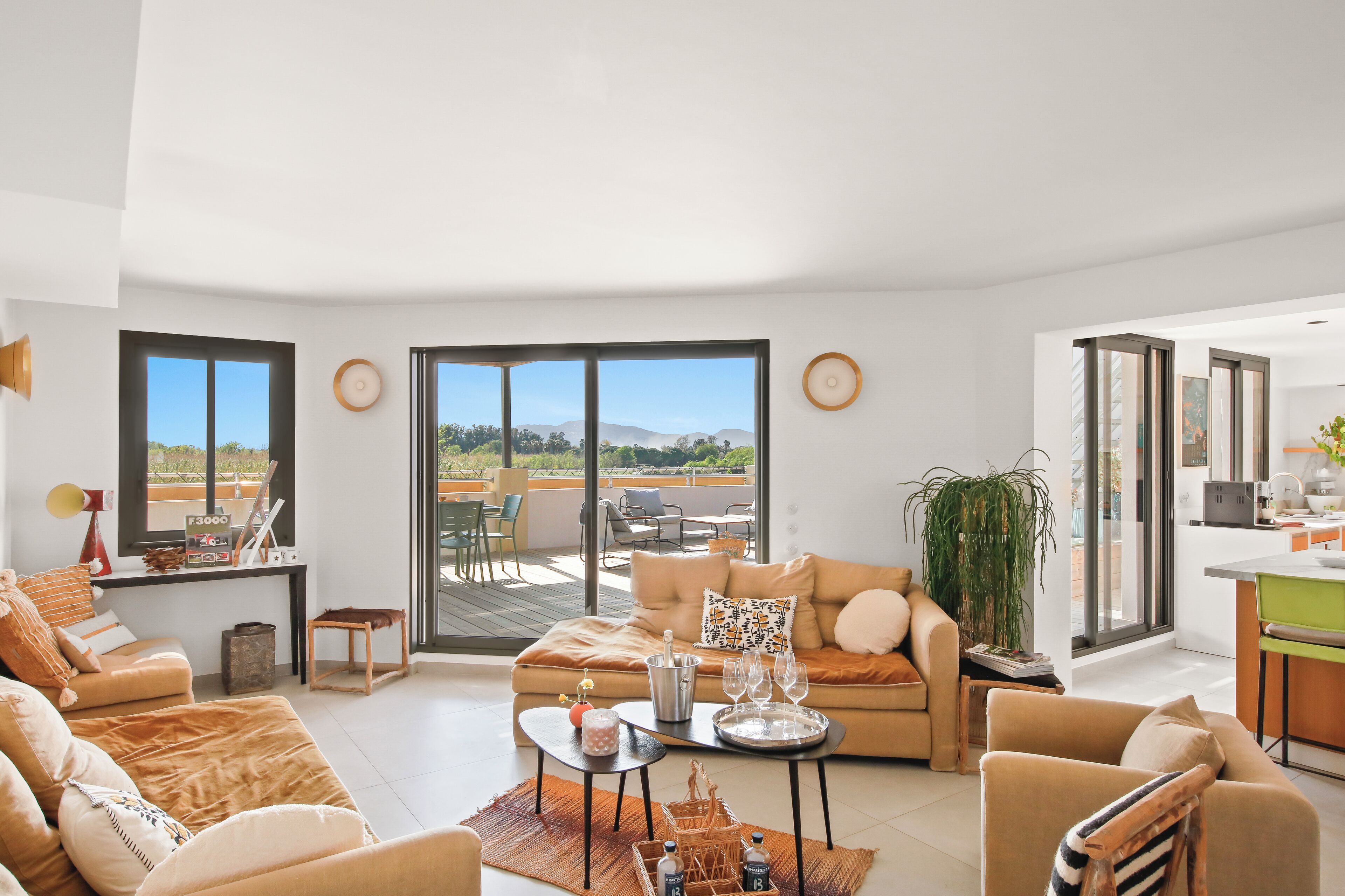 A spacious living area with a sofa, a coffee table, and a dining table. There is a balcony with outdoor furniture and a view of the landscape.