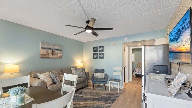 A living room with a ceiling fan, a dining table with chairs, a sofa, a TV, and a refrigerator. - Seaside Escape: Beautifully Appointed 1 BR Condo (North Wildwood)