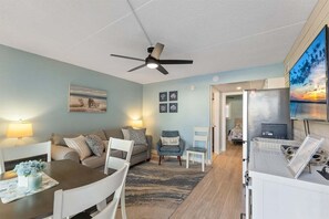 A living room with a ceiling fan, a dining table with chairs, a sofa, a TV, and a refrigerator.