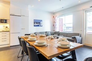 Une cuisine moderne avec un coin repas, des meubles blancs, une table en bois dressée pour six personnes et un canapé gris.