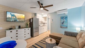 A modern living room with a sofa, a TV, a ceiling fan, and a kitchen in the background. - Gorgeous 2 Bedroom Condo with Two Heated Pools (North Wildwood)
