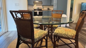 Een eetruimte met een tafel met glazen blad, omringd door rieten stoelen met bloemenkussentjes.