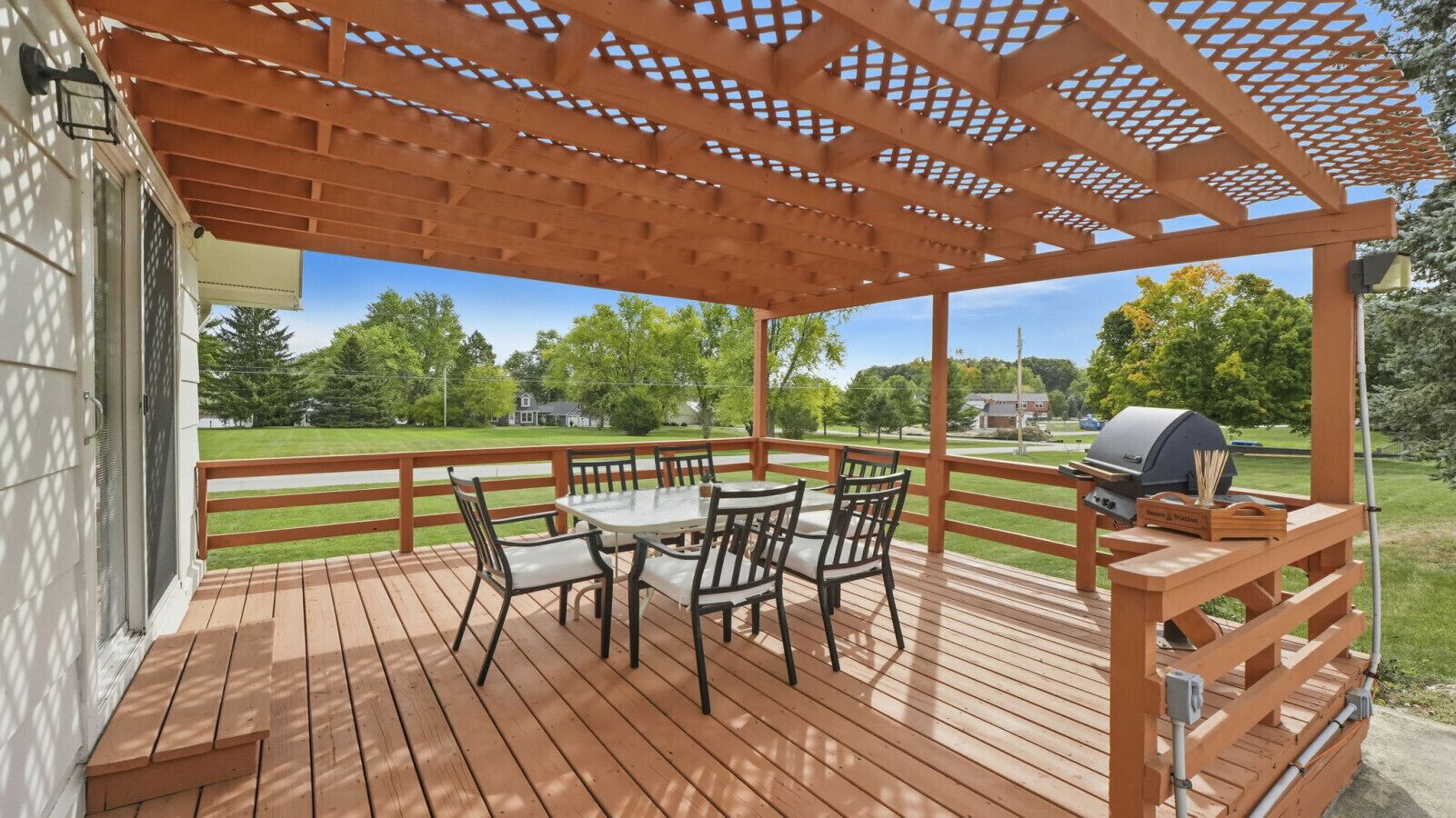 Una terraza de madera con pérgola, mesa y sillas de exterior, y una parrilla.