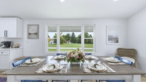A dining room with a large table set for six, white cabinets, and a view of a green landscape through large windows.