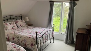 A bedroom with two beds, a window with curtains, and a small wooden table. - Rose Cottage (Hamilton)