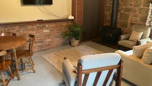 A cozy living room with a brick fireplace, a stone wall, a flat-screen TV, a dining table with chairs, and a comfortable sofa. - Rose Cottage (Hamilton)