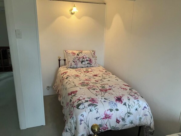 Una habitación pequeña con cama individual, colcha de flores, lámpara de noche y luz en la pared.